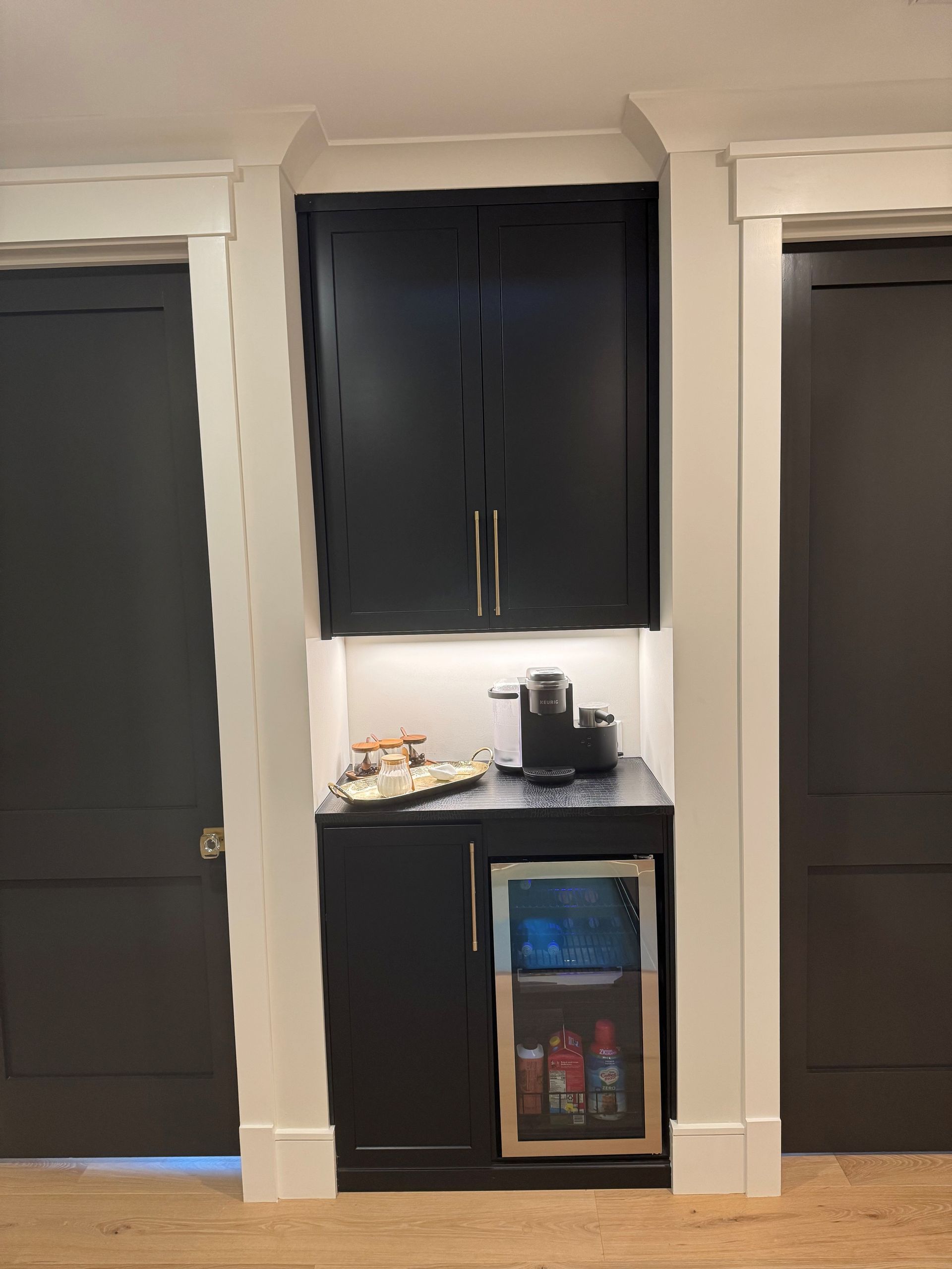 Black built-in coffee station with upper cabinets, lower cabinet with a beverage cooler, and under-cabinet lighting.