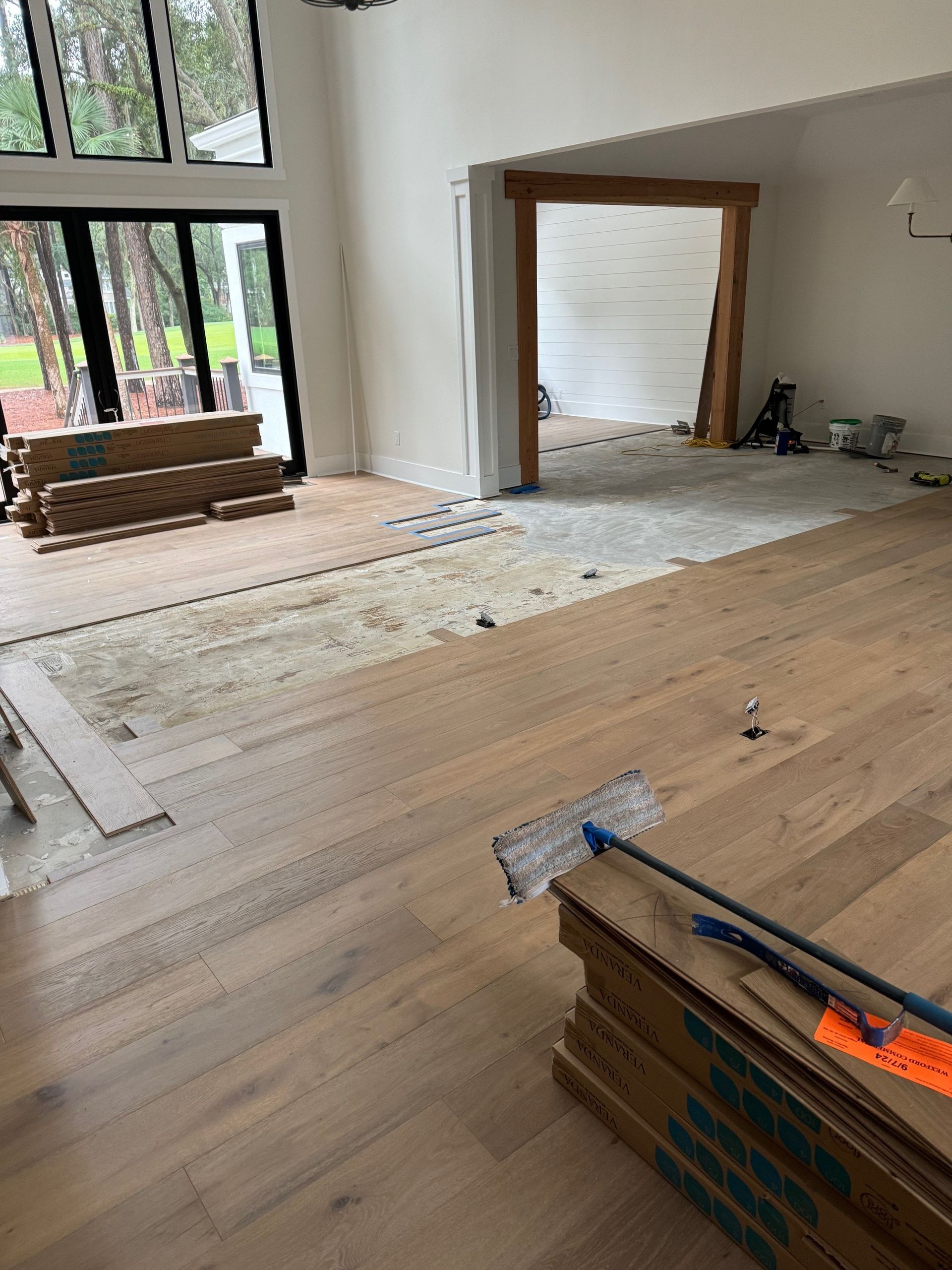 Installing hardwood flooring in a room with large windows and a doorway. Boards are in place and stacked.