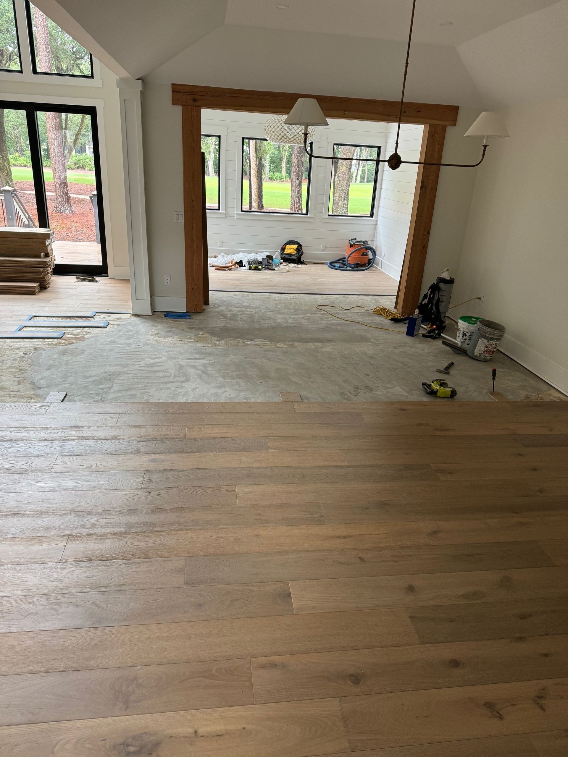 Interior view of a room under construction with new wooden flooring. A doorway leads to a windowed area with tools.