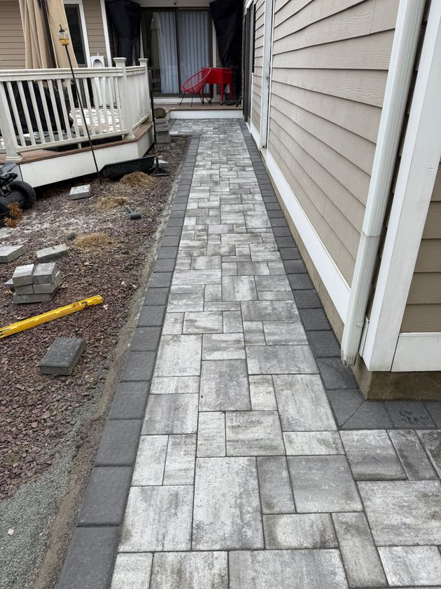 A newly installed stone paver walkway runs alongside a house with beige siding, featuring a gray rectangular border.