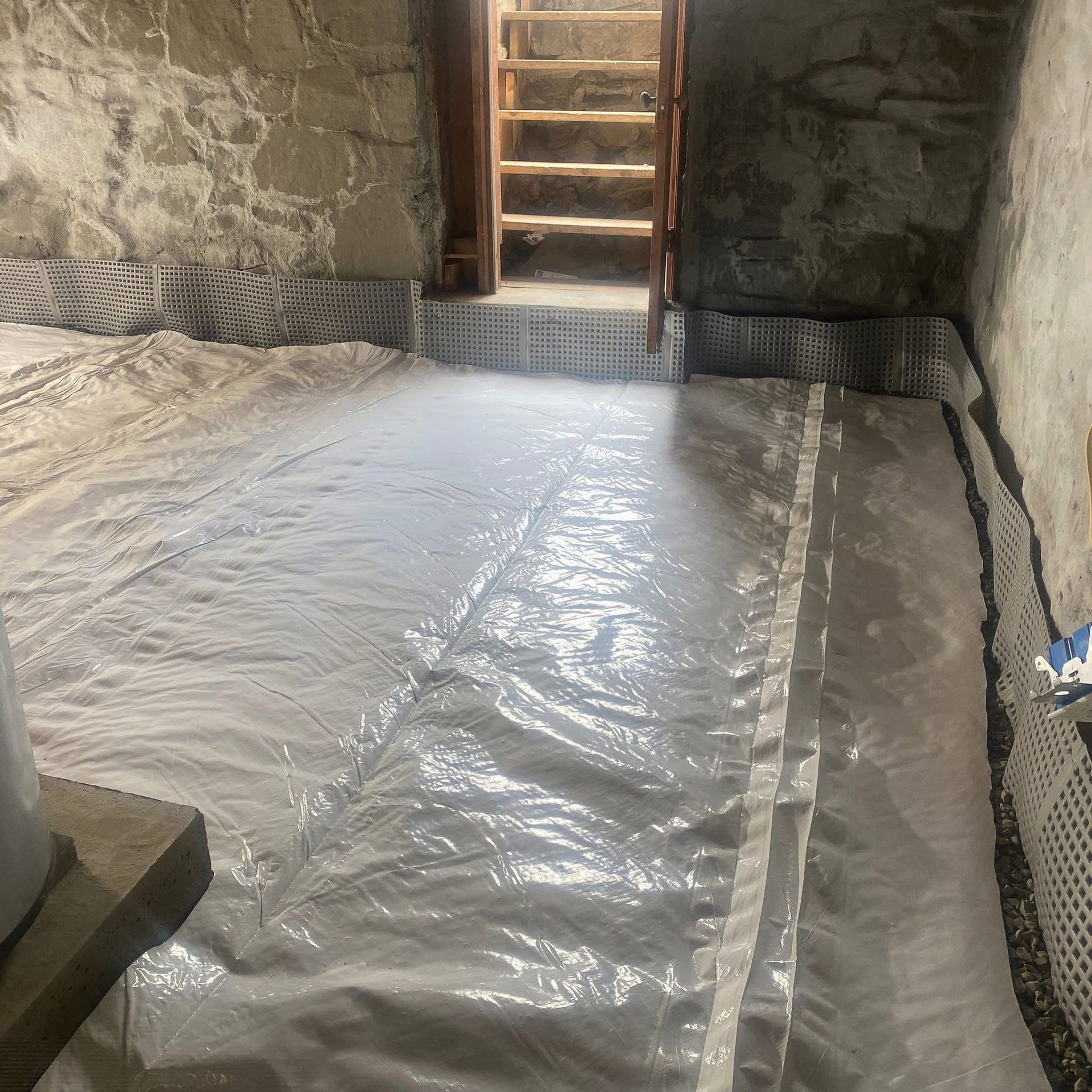 A crawlspace with stone walls and a floor covered by a thick white plastic vapor barrier, leading to a wooden staircase.