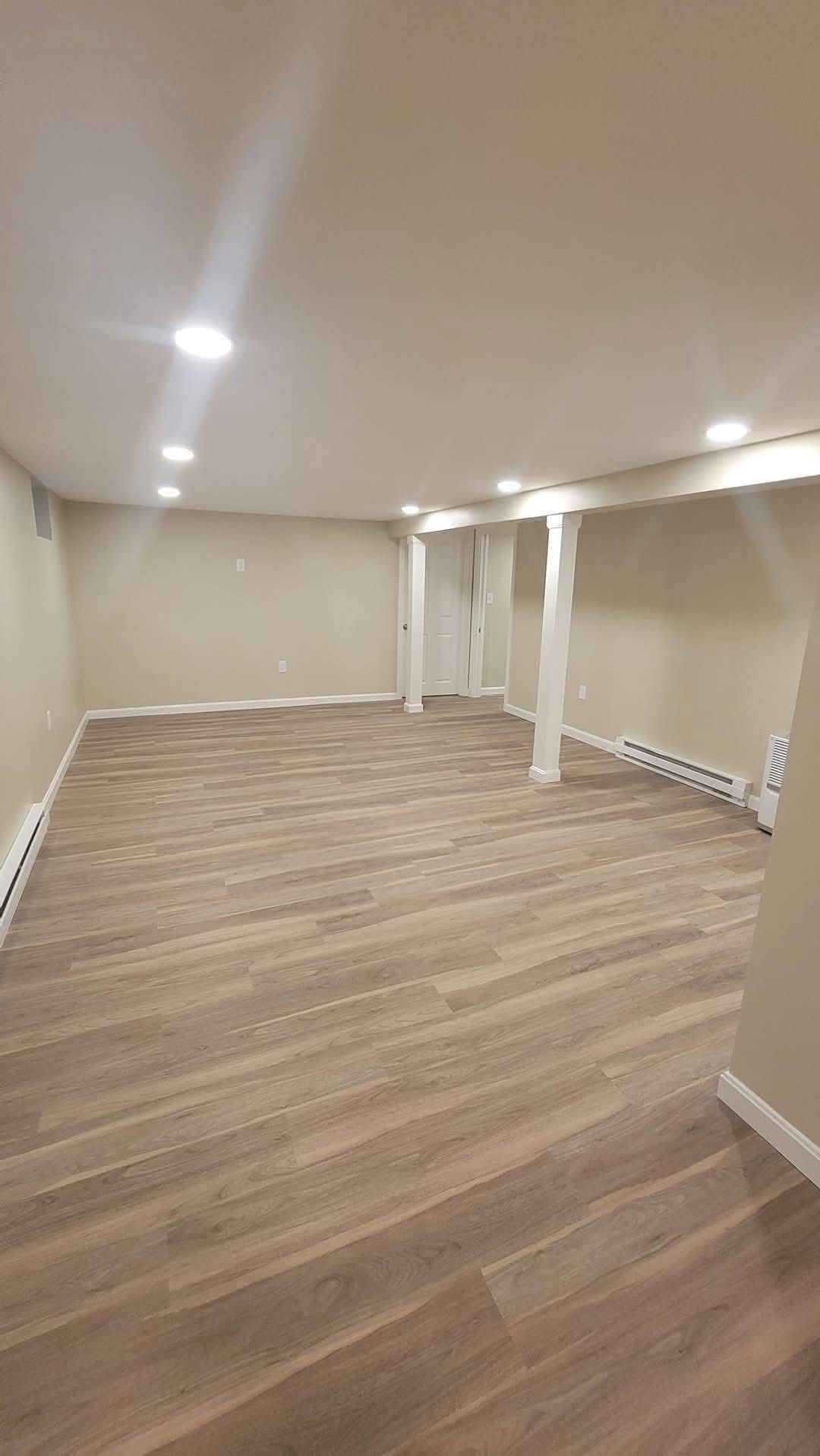 A spacious basement with light beige walls, wood-look flooring, recessed ceiling lights, and two white support columns.