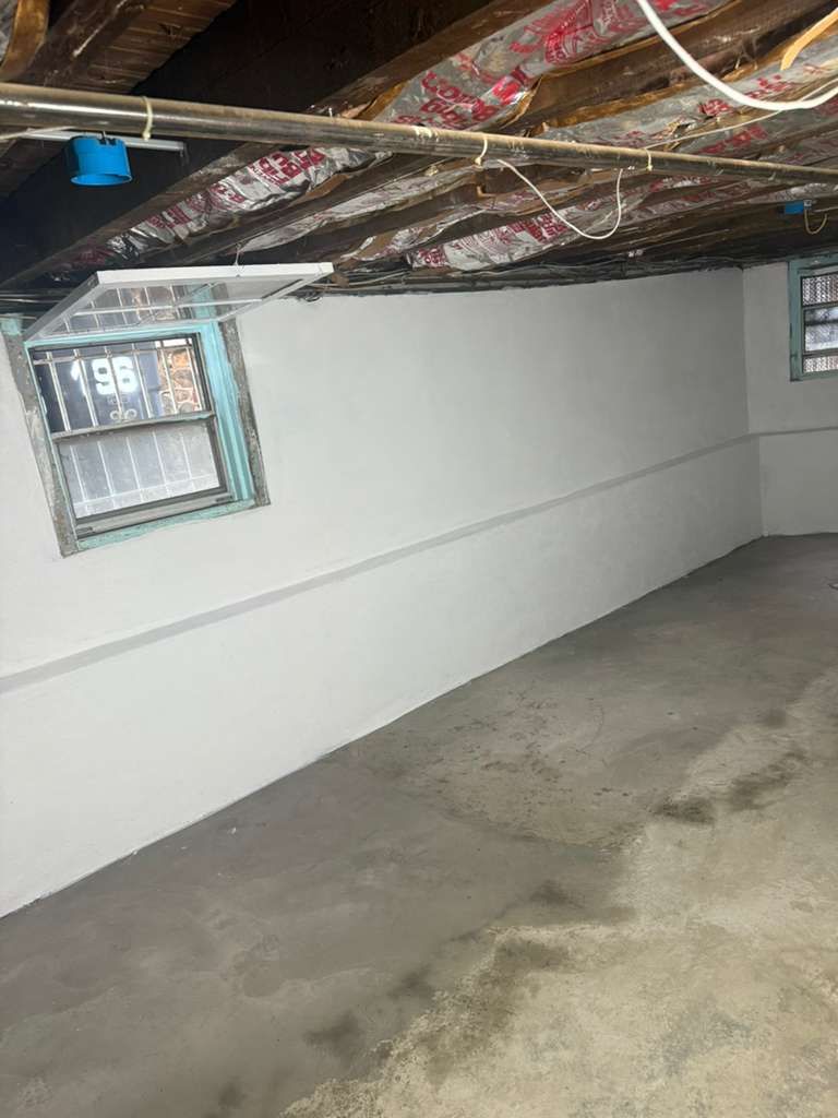 A basement corner featuring white concrete walls, a small window with a blue frame, and exposed floor joists above.