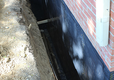 A view of a trench dug along a brick house foundation, which is coated in black waterproofing sealant.
