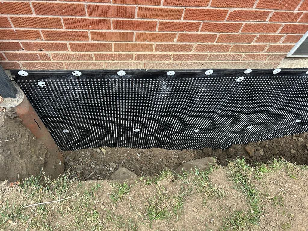 Black foundation drainage membrane installed against a brick wall above an excavated trench in the ground.
