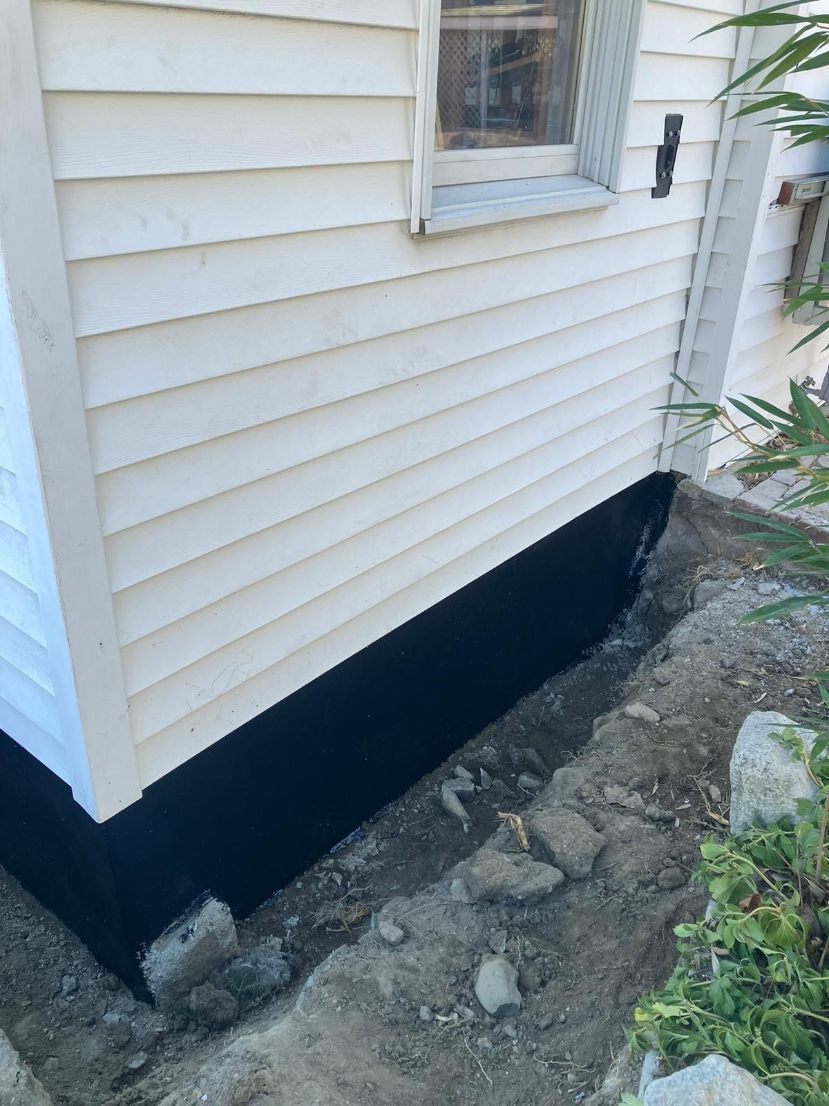 Exterior wall of a house with light-colored siding above a freshly applied black waterproof foundation coating.