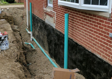 A trench dug along a brick house foundation, showing black waterproof coating and newly installed green drainage pipes.