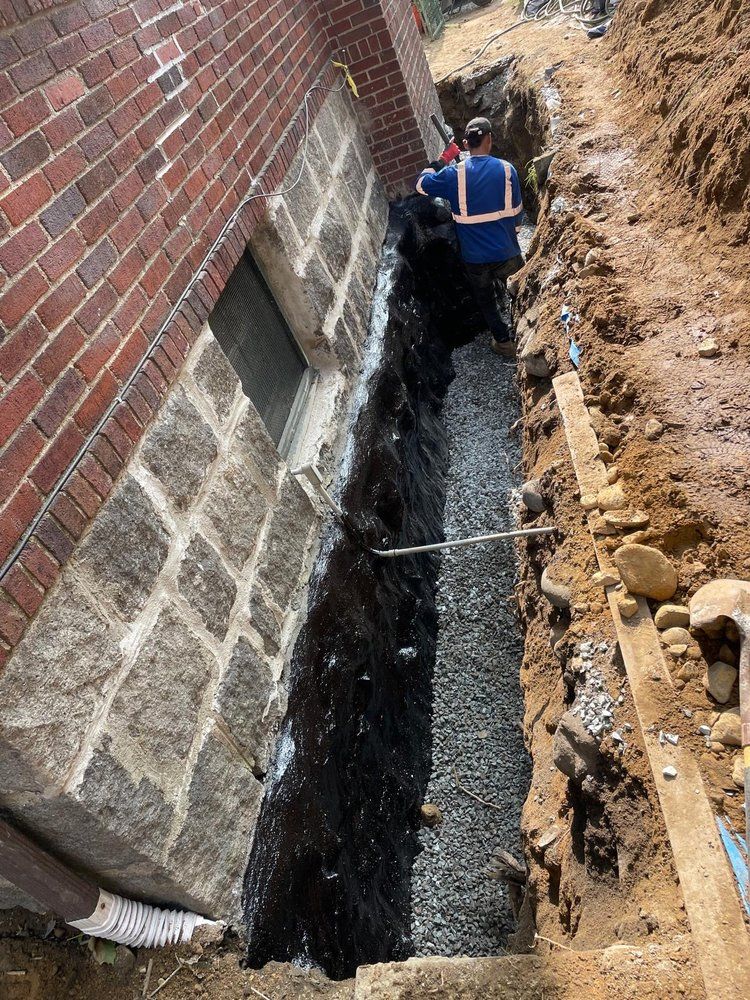 A construction worker in a blue vest works in a narrow trench beside a brick and stone building foundation with drainage.