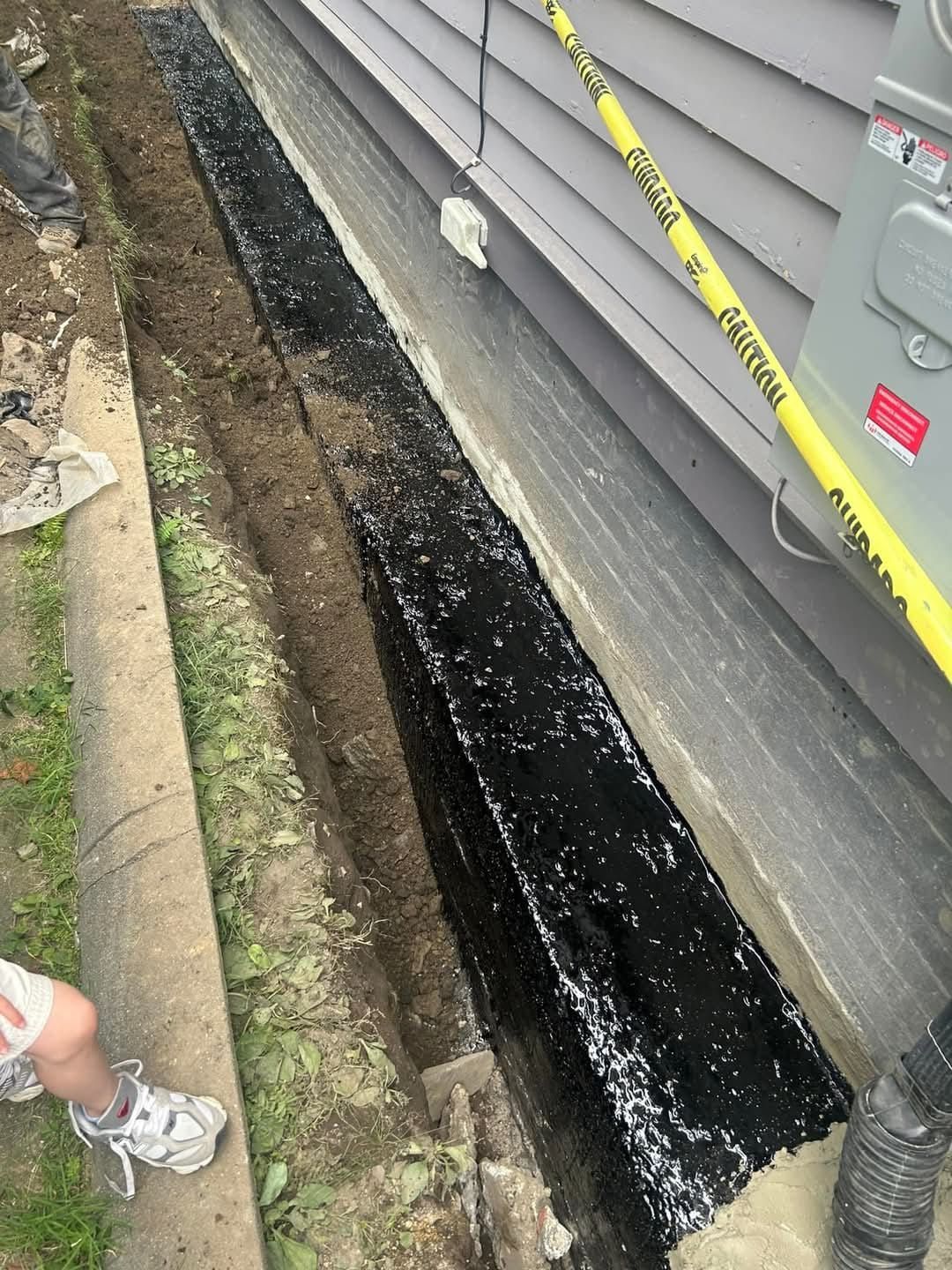 A foundation trench along a house wall, coated with a black waterproof sealant, with caution tape and a drainage pipe.