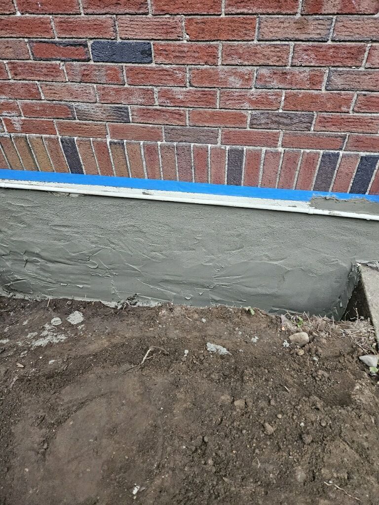 A freshly applied layer of gray concrete parging on a house foundation wall below red brick siding and blue painter's tape.