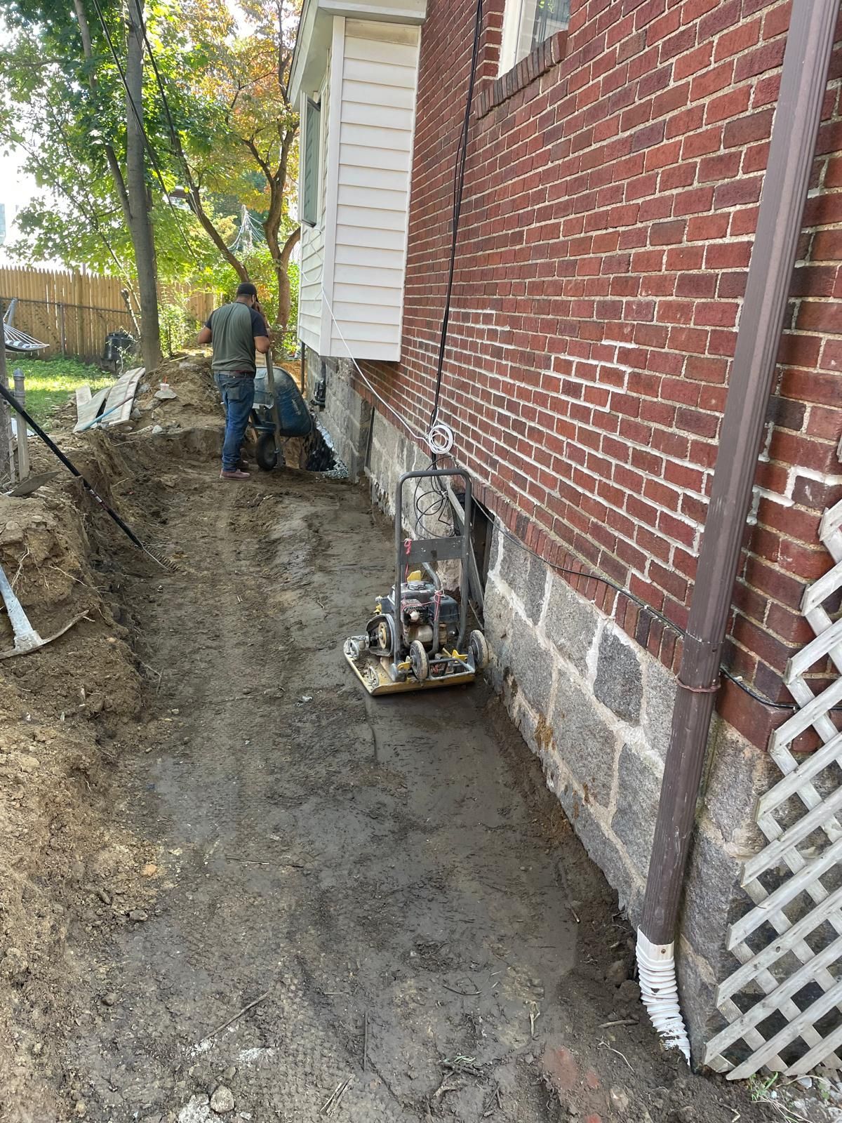 A worker operates a plate compactor on dirt next to a brick building's foundation and a white exterior wall.