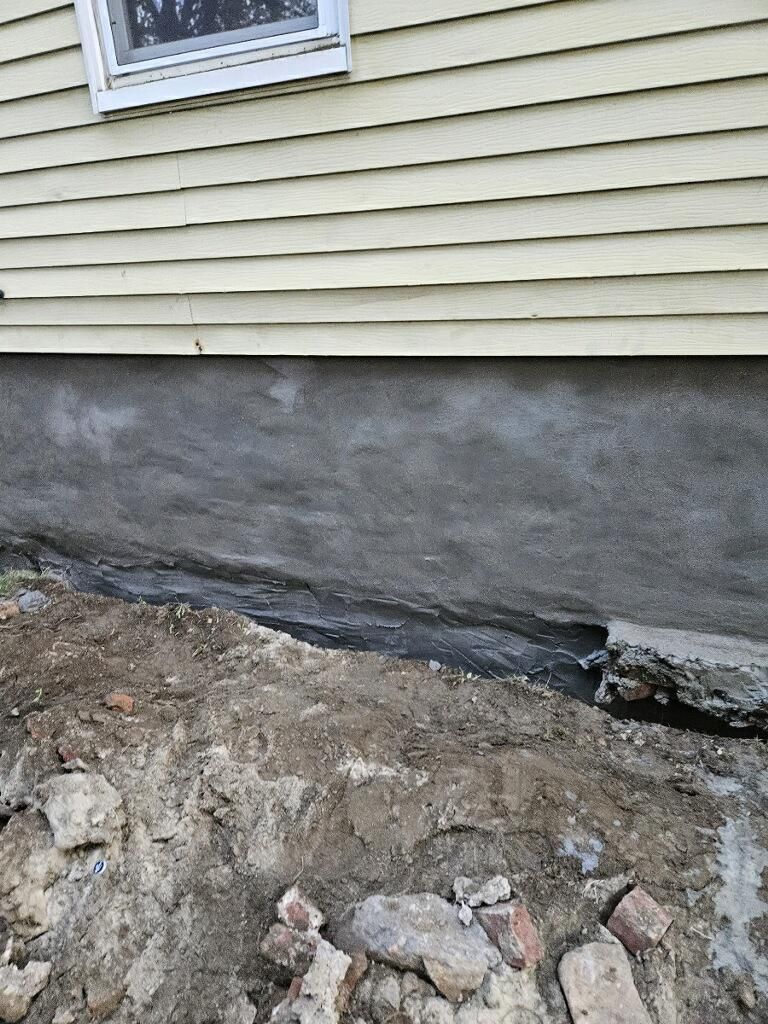 Light yellow siding sits above a freshly parged grey concrete foundation, with disturbed soil and debris at its base.