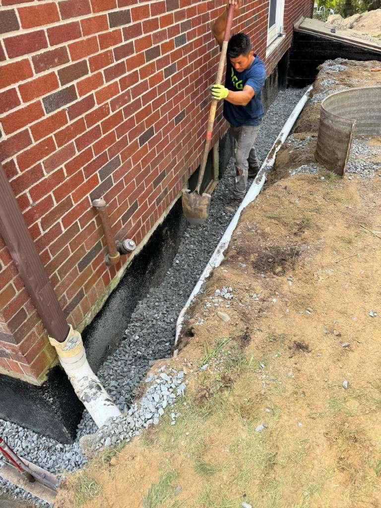 A person in a blue shirt uses a shovel to fill a foundation drainage trench along a brick house with gravel.