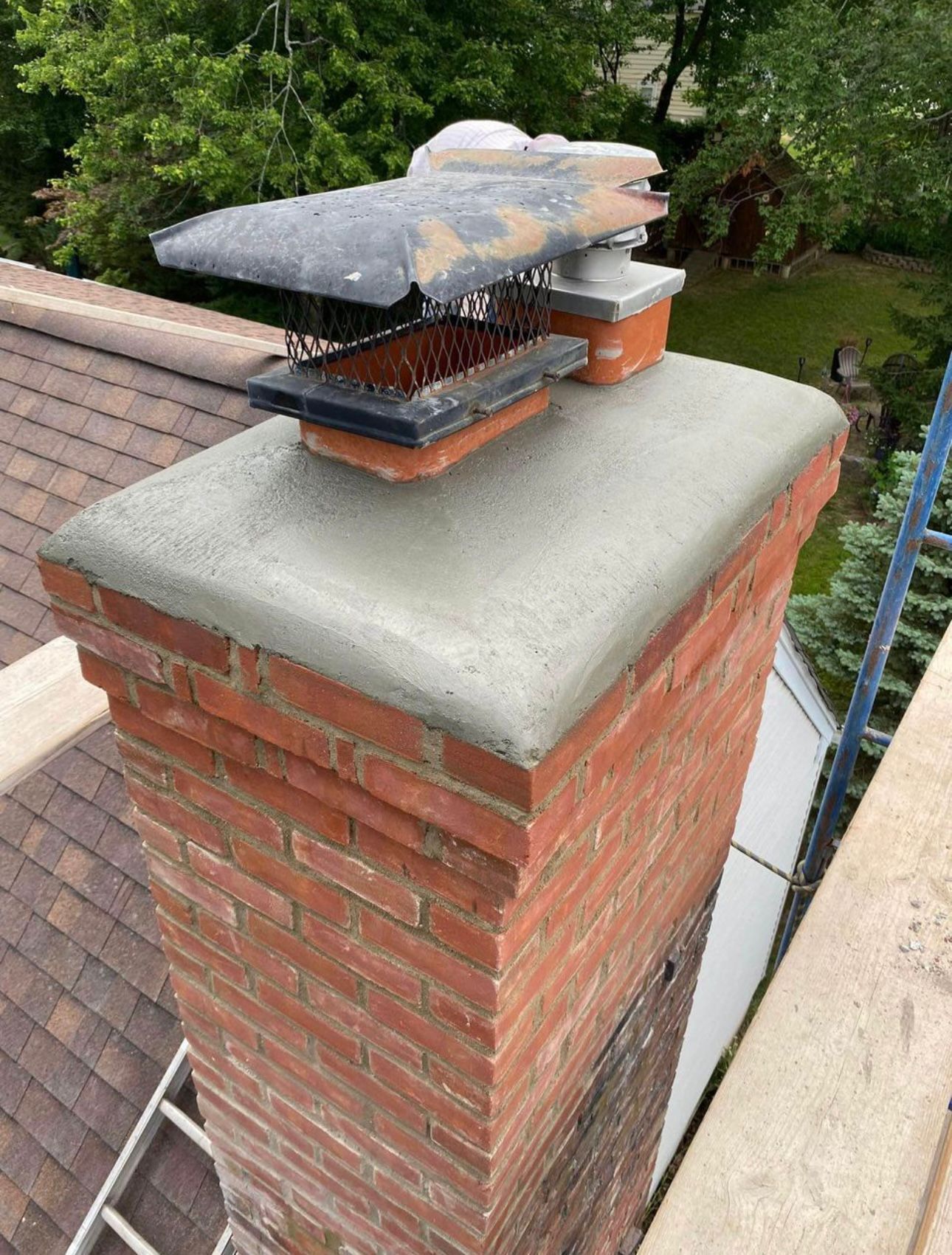 A newly poured concrete chimney crown sits atop a brick chimney with a metal cap and flue vent on a shingled roof.