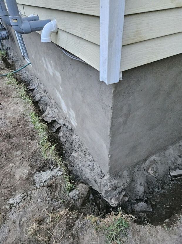 A gray concrete building foundation meets beige siding, showing exposed footing in a muddy trench alongside the house.