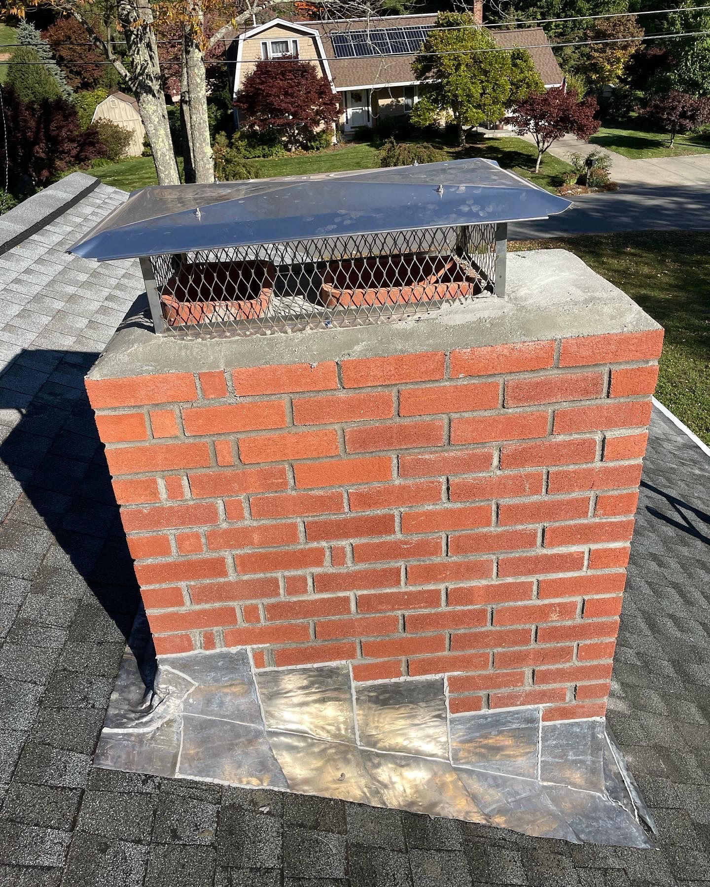 A red brick chimney with a metal cap and wire mesh, centered on a shingled residential roof.