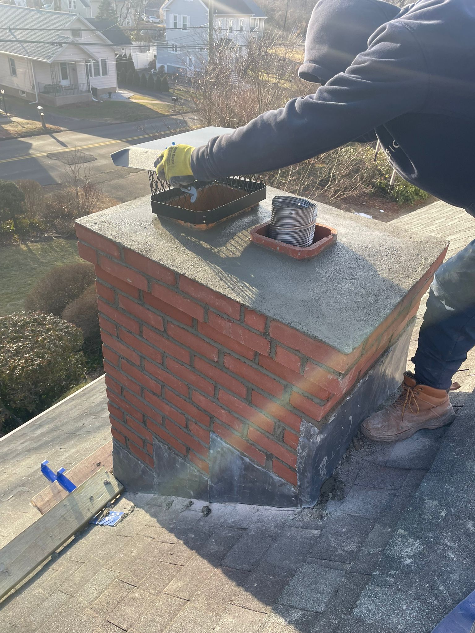 A person in a dark hoodie and work boots seals the mortar of a brick chimney on a shingled residential roof.