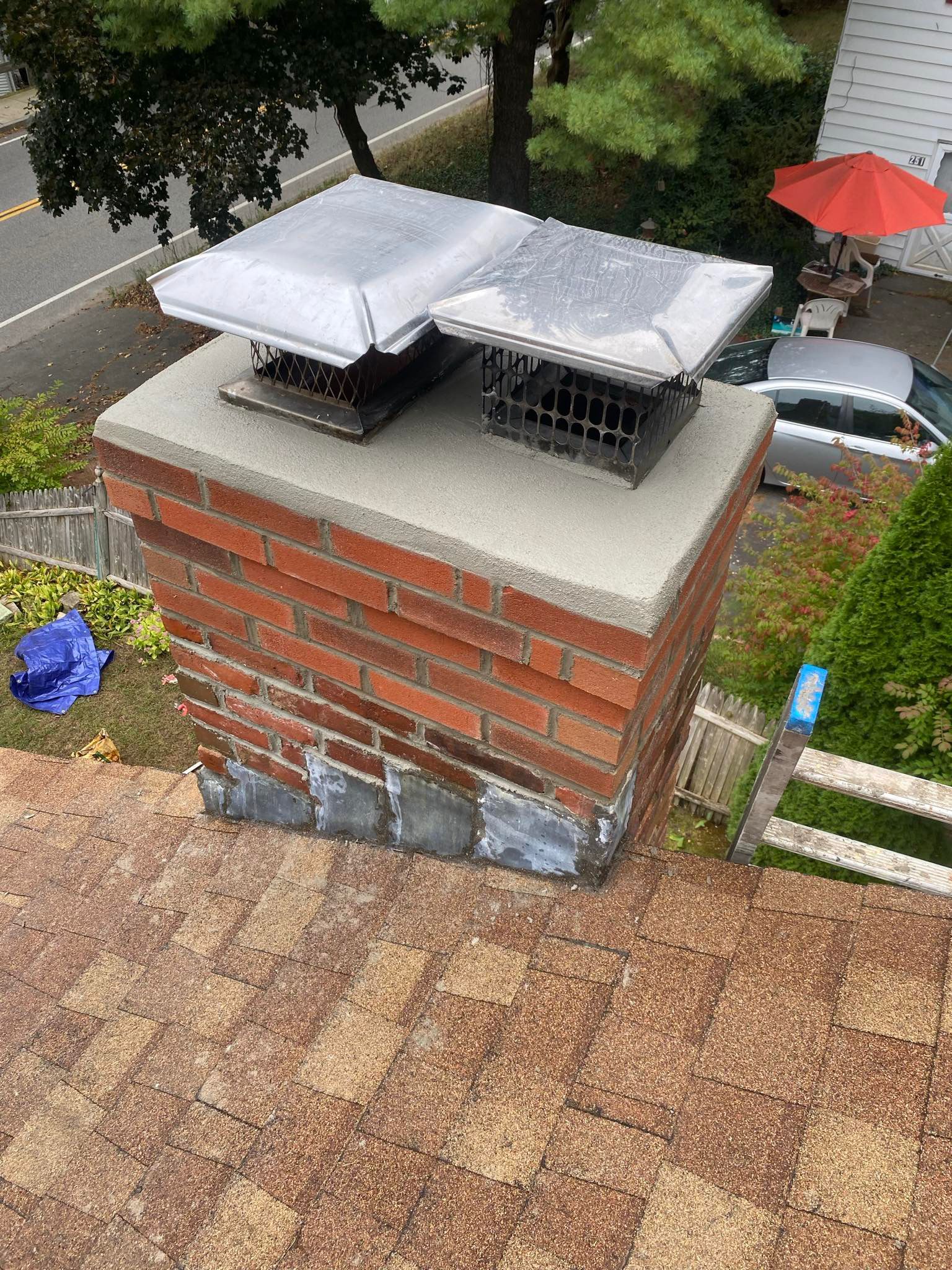 A brick chimney on a shingled roof features a new concrete crown and two stainless steel chimney caps.