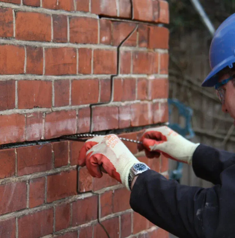 A construction worker in a blue hard hat inserts a reinforcement bar into a crack in a brick wall for structural repair.