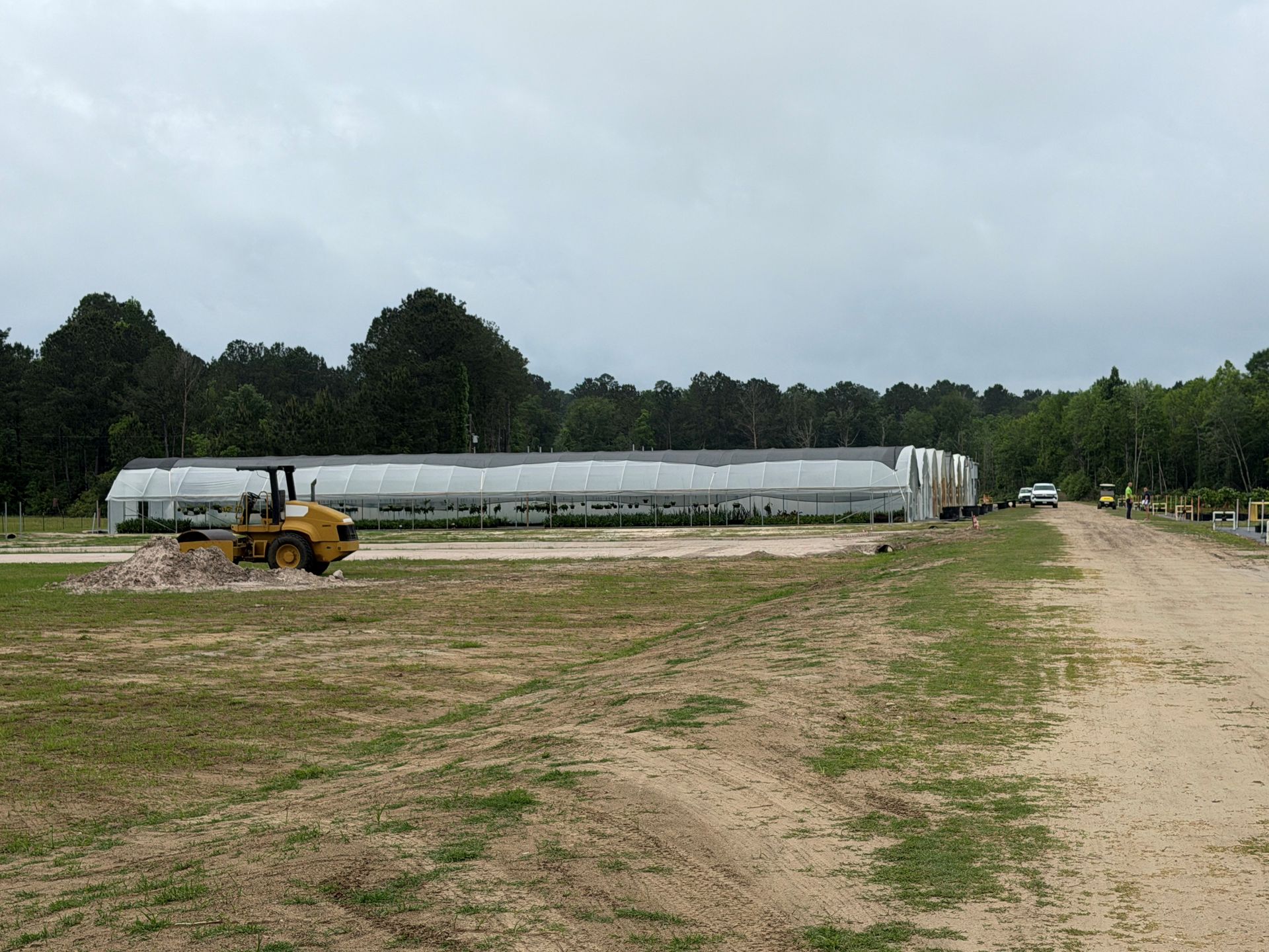 Yellow tractor near long silver greenhouse in a dirt field under cloudy sky