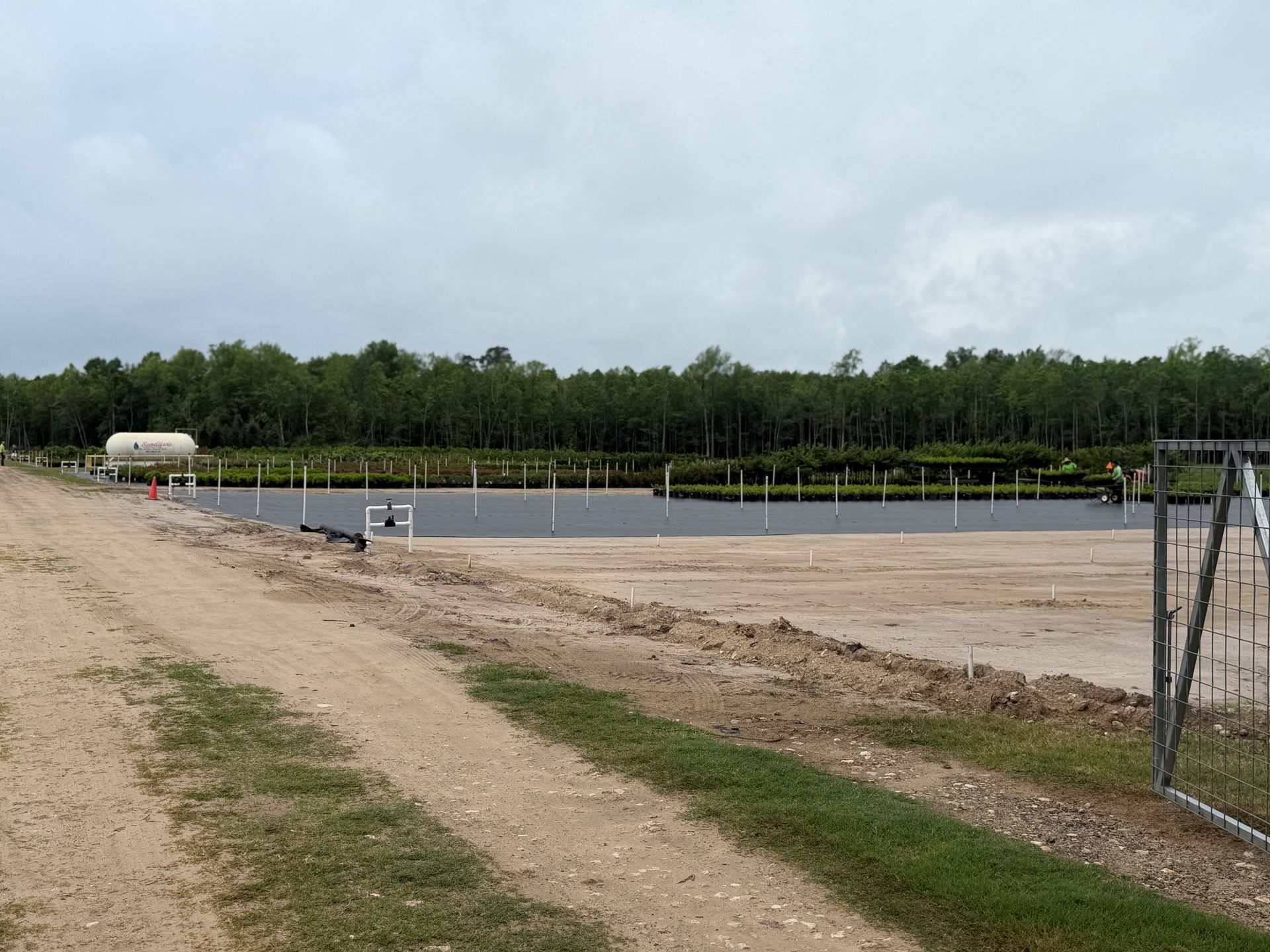 Gravel road beside a fenced gravel lot under a cloudy sky, with trees in the background.