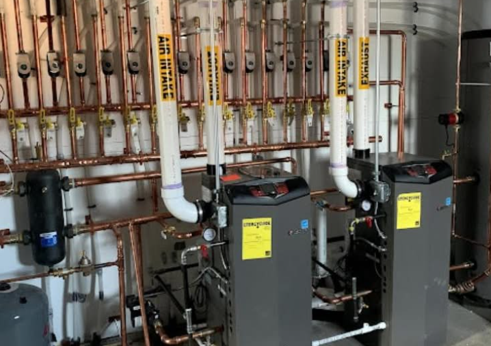 Two gray water heaters with copper pipes in a utility room. Yellow warning stickers visible.