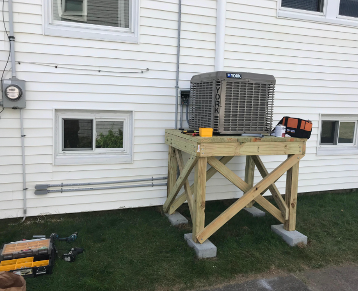 A York air conditioner sits on a wooden stand next to a white house with gray electrical components.