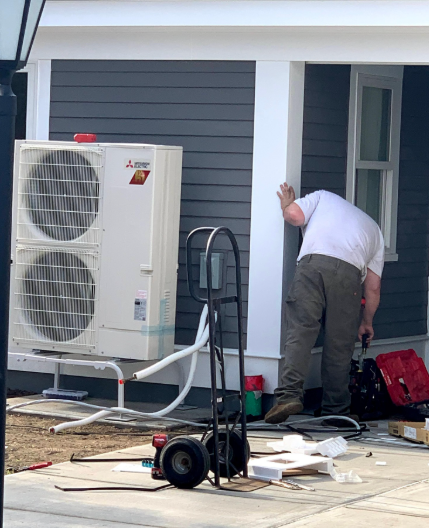 Man working on a building's exterior near a white HVAC unit; equipment scattered on the ground.