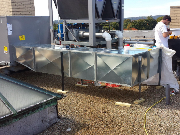 HVAC equipment on a rooftop; a person works nearby. Silver ductwork, cooling tower, and a grey unit on a rooftop.