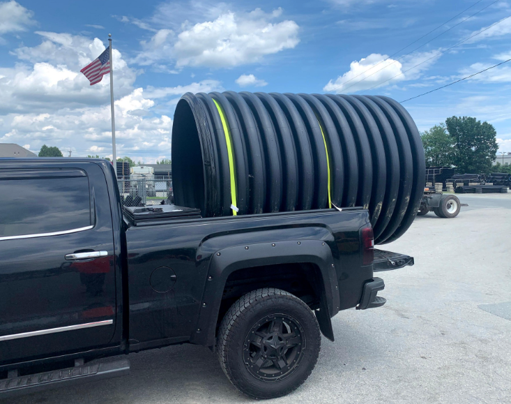 Black pickup truck carrying large, black corrugated plastic pipe, secured with straps. American flag in background.