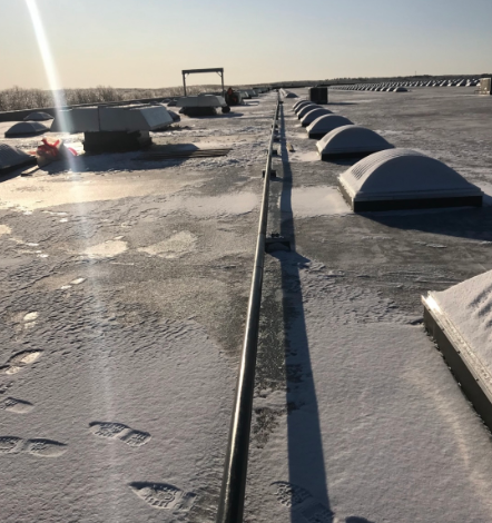 A snow-covered flat roof with skylights, vents, and a metal safety rail under a sunny sky.