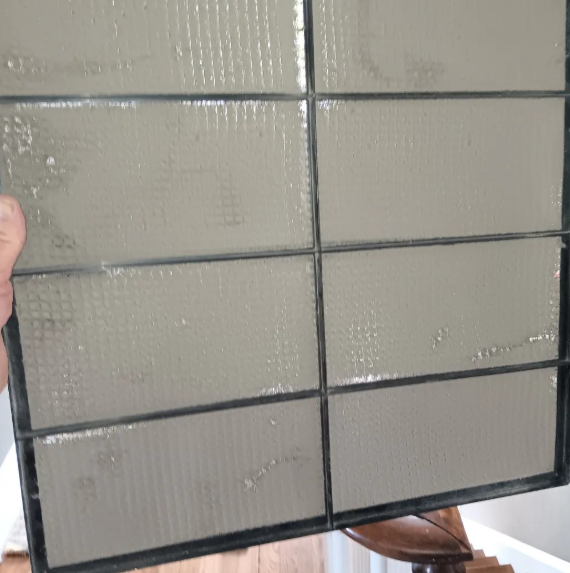 Dirty air filter held by a person; rectangular, with dark grid lines, coated in gray grime.