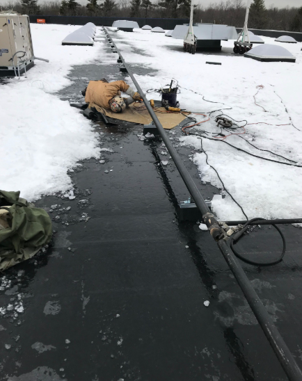 Person working on a snowy rooftop, with equipment and a long black pipe visible.