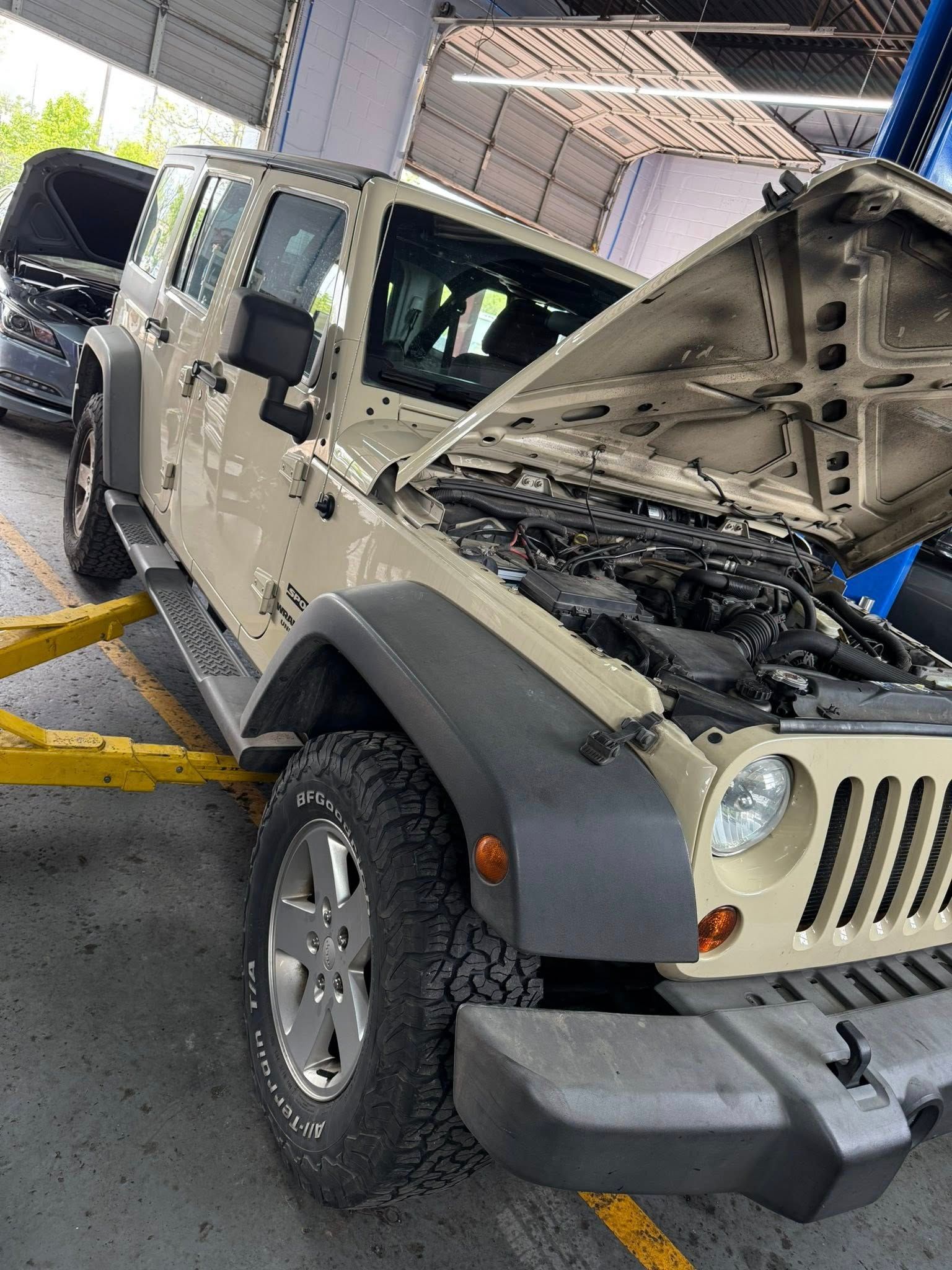 Beige Jeep Wrangler with hood open in a garage, showing the engine compartment.