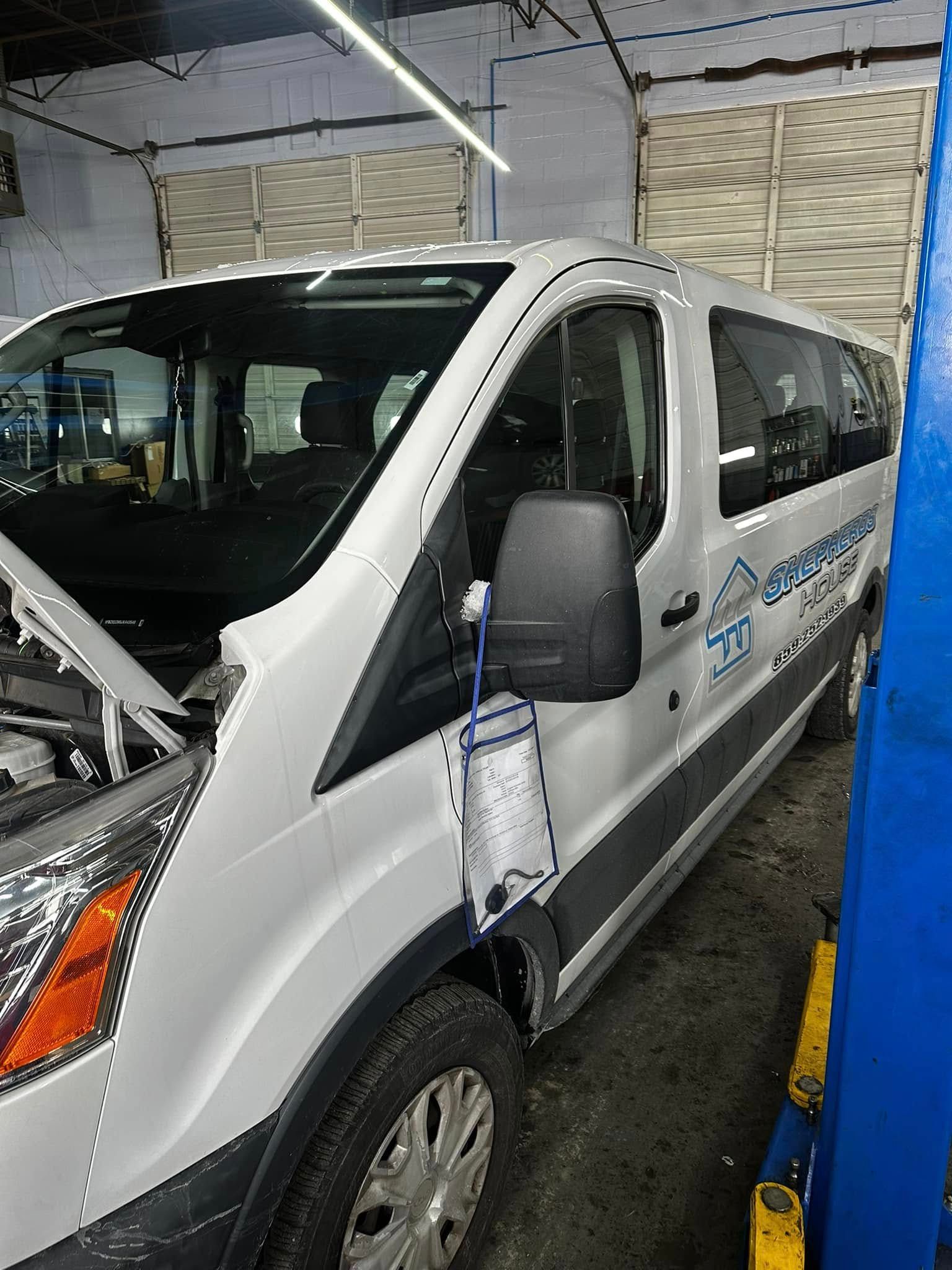 White van in an auto shop bay, with a front hood open and a blue lift post beside it.