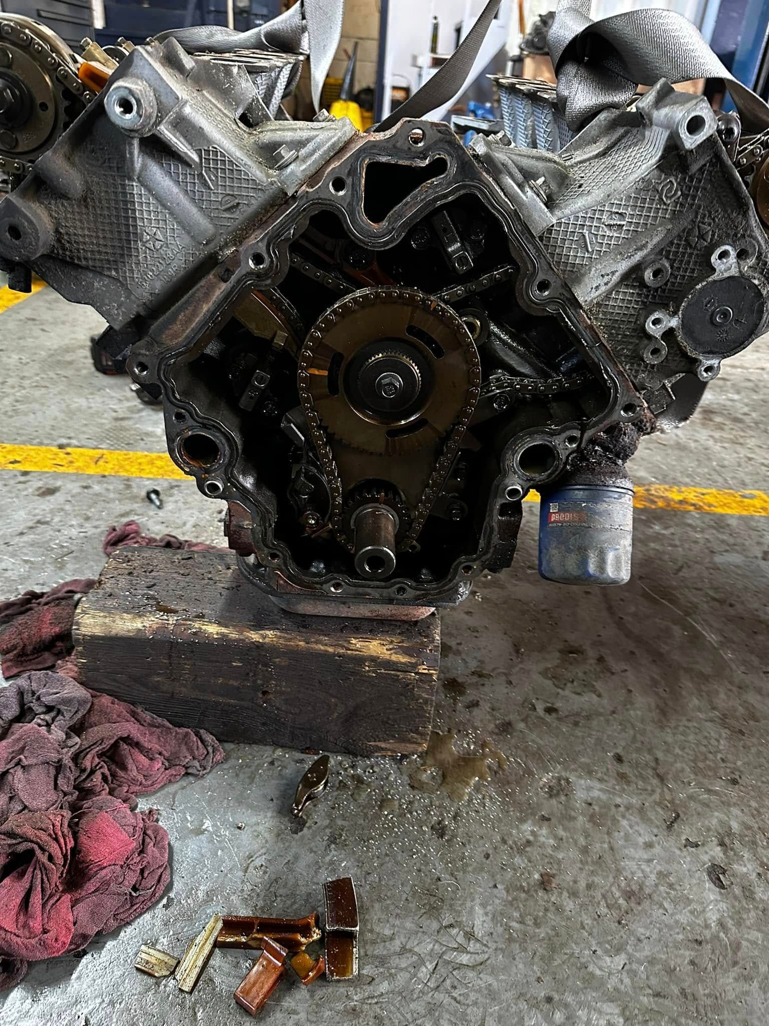 Open car engine on the floor with a timing chain and tools nearby, beneath a vehicle chassis.
