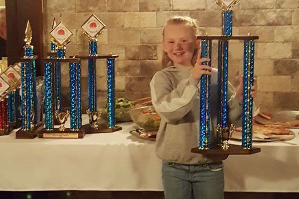 A young girl is holding a trophy in front of a table full of trophies.