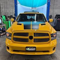 A yellow dodge ram truck with a blue stripe on the hood is parked in a garage.