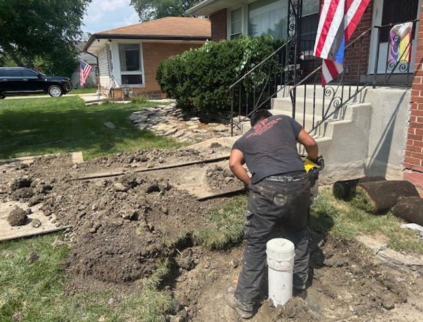 A man digs near a white pipe outside a house. Soil is piled up, and a roll of sod sits nearby.