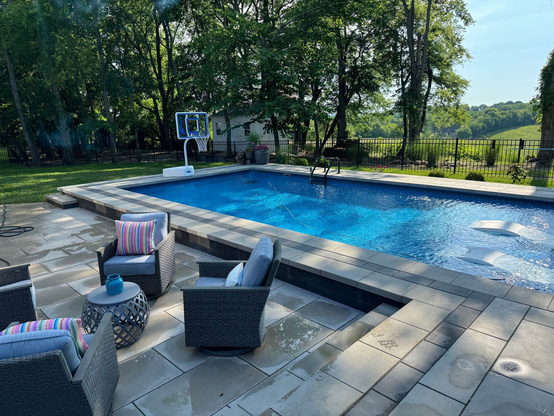 A large swimming pool is surrounded by a patio with chairs and a table.