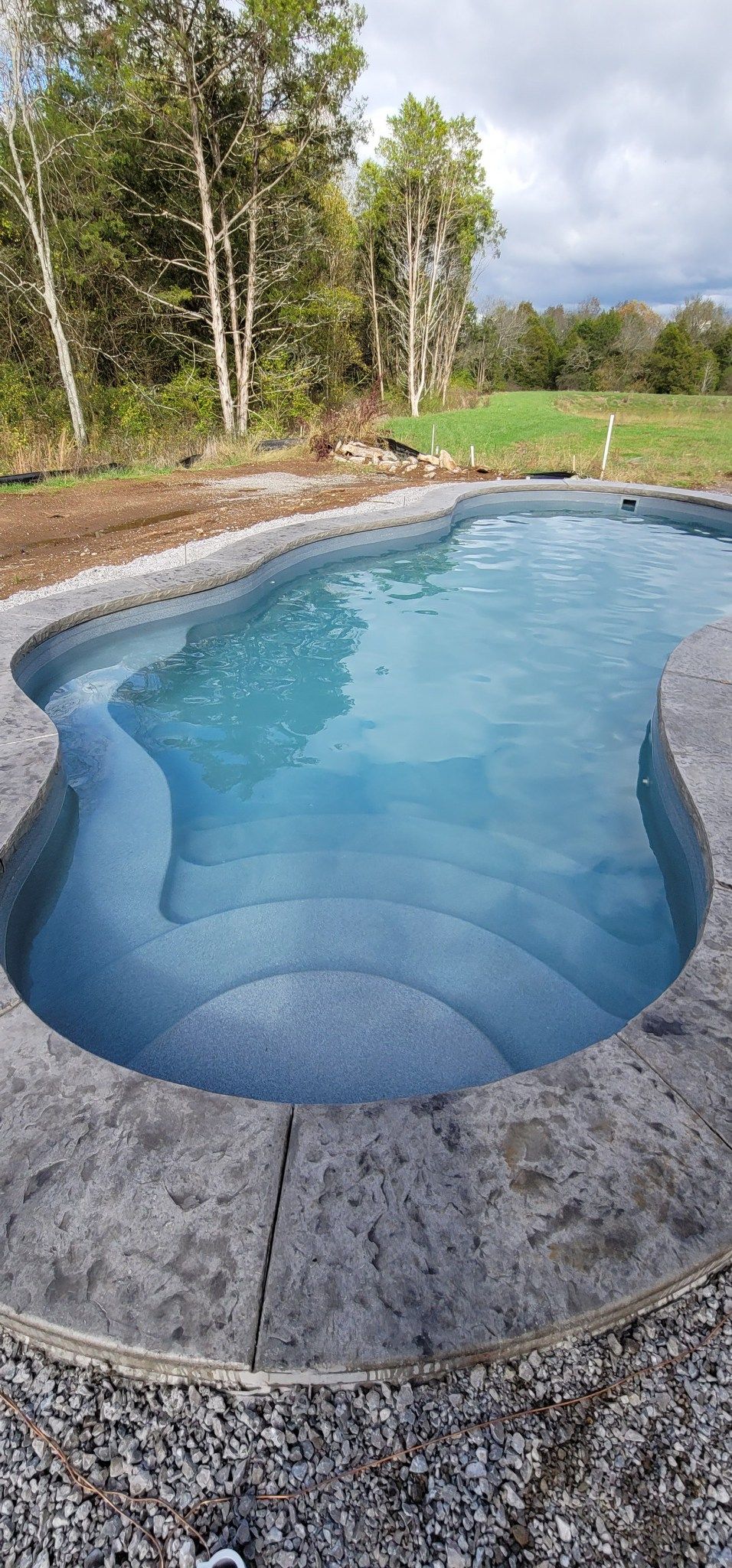 A large swimming pool is surrounded by gravel and trees.