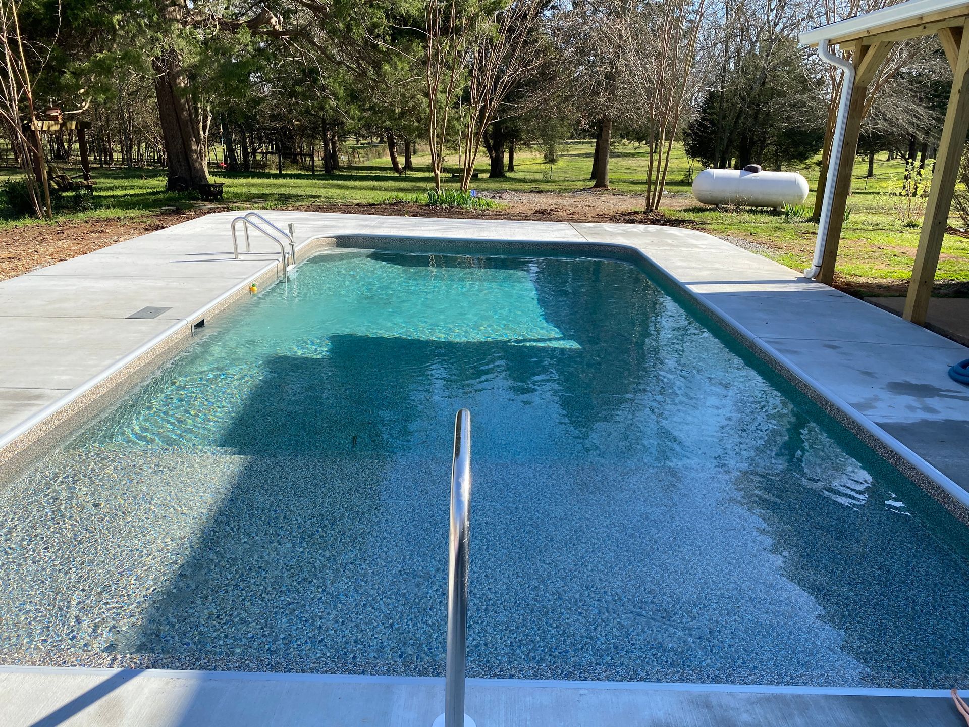 A large swimming pool is surrounded by trees and a propane tank.