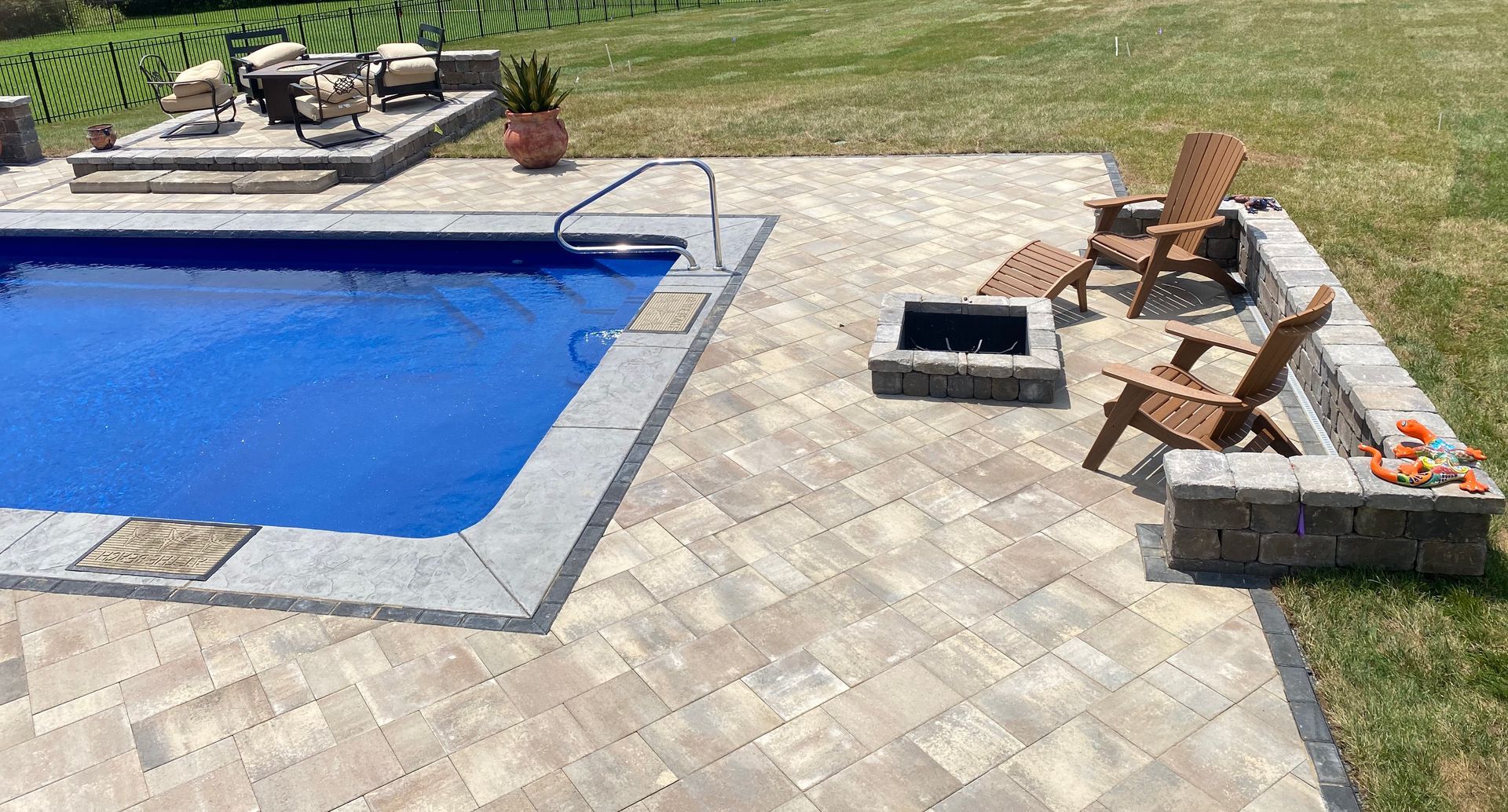 A large swimming pool is surrounded by a patio with chairs and a fire pit.