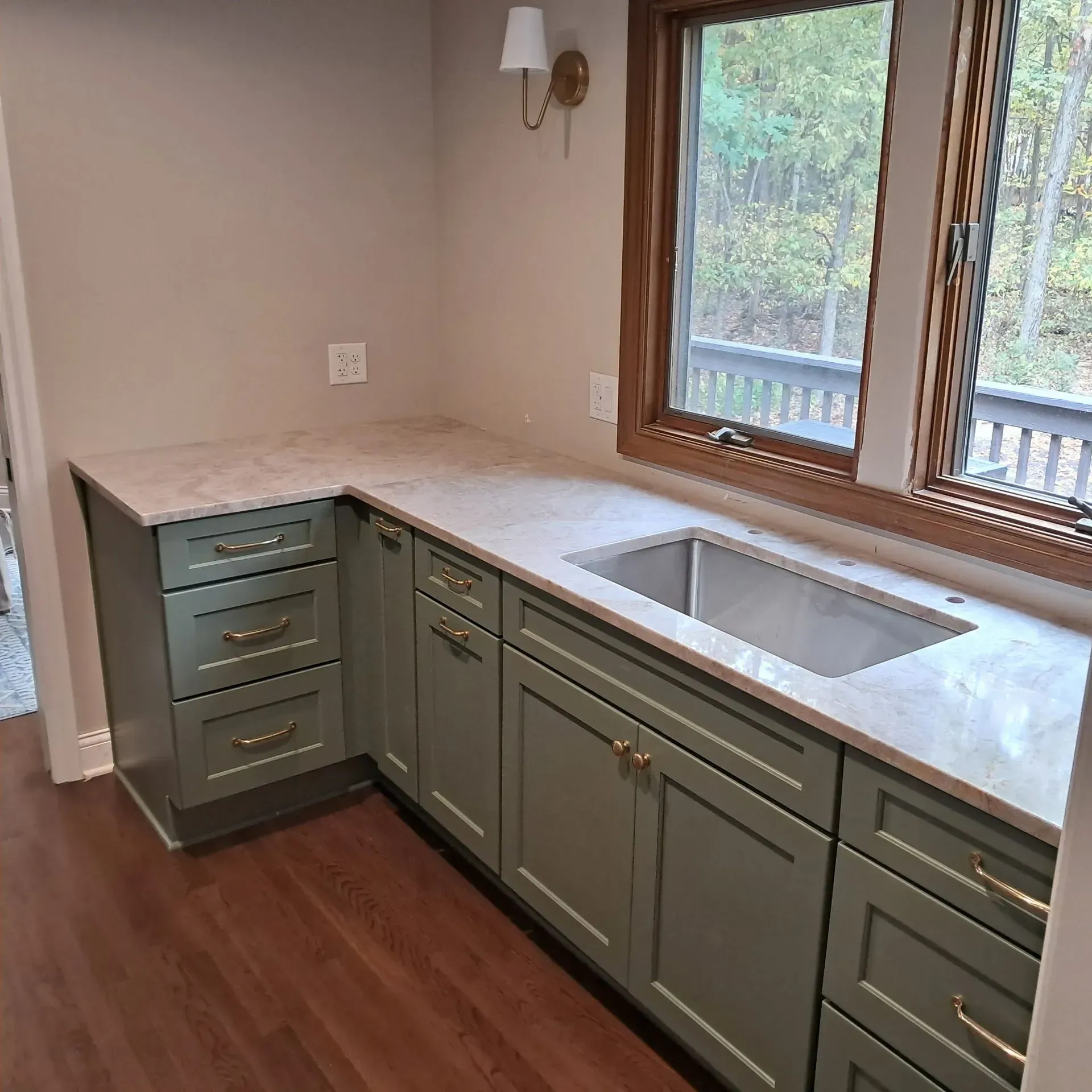 Green kitchen cabinets with a light countertop, sink, and window overlooking a deck and trees.