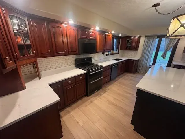Cherry wood kitchen cabinets with white countertops, black appliances, and light wood flooring.