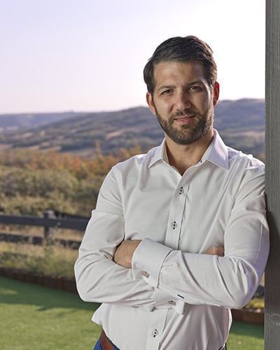 A man with a beard is leaning against a pole with his arms crossed.