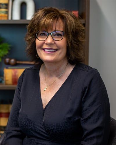 A woman wearing glasses and a black shirt is sitting in front of a bookshelf.