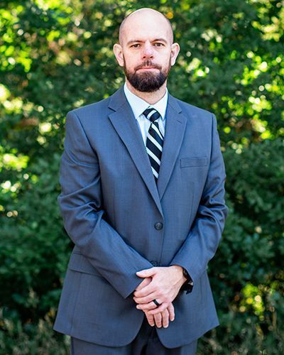 A man in a suit and tie is standing with his hands folded in front of trees.