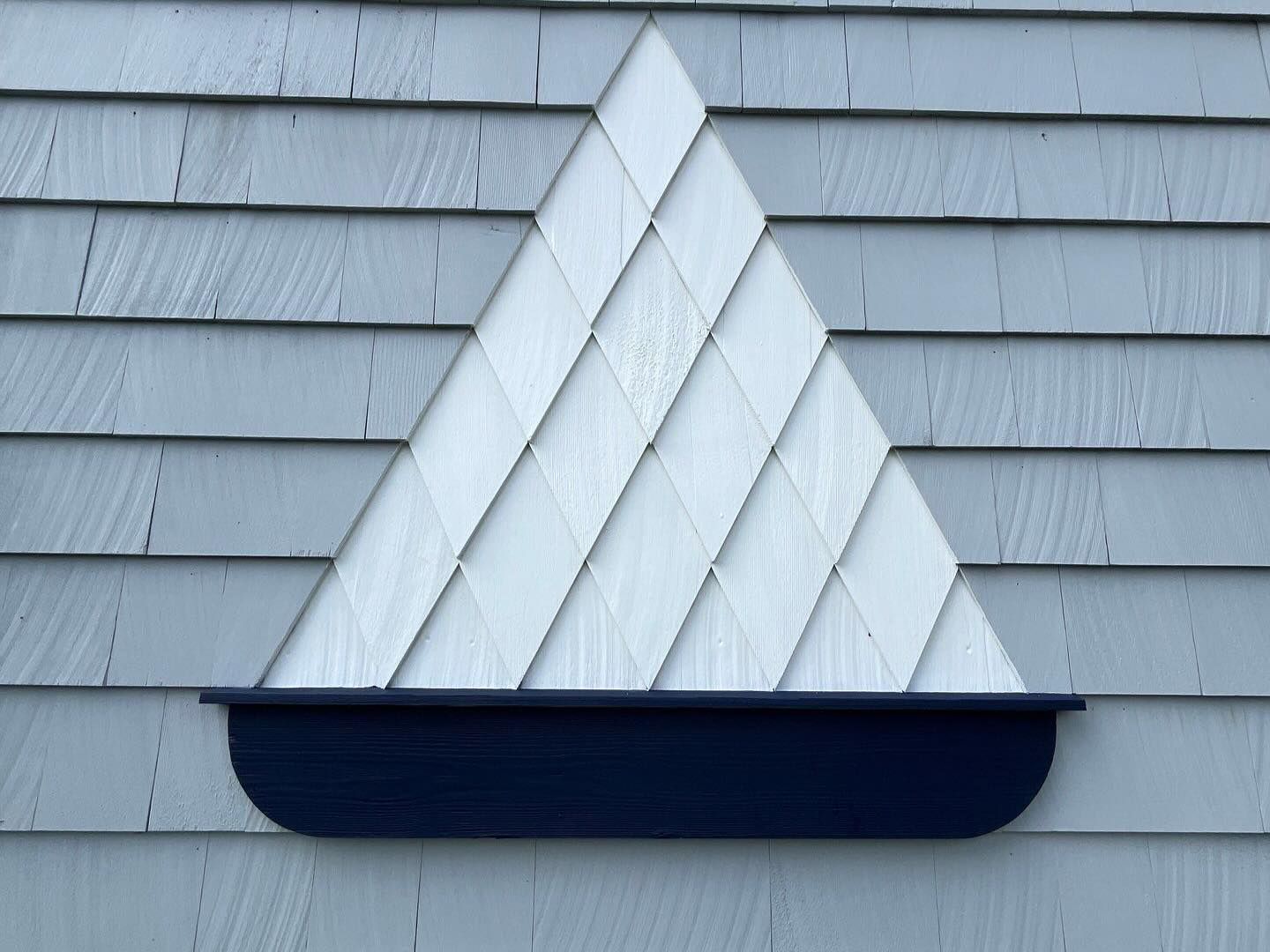 Blue and white shingle design in the shape of a triangle above a dark blue planter on a gray shingled wall.