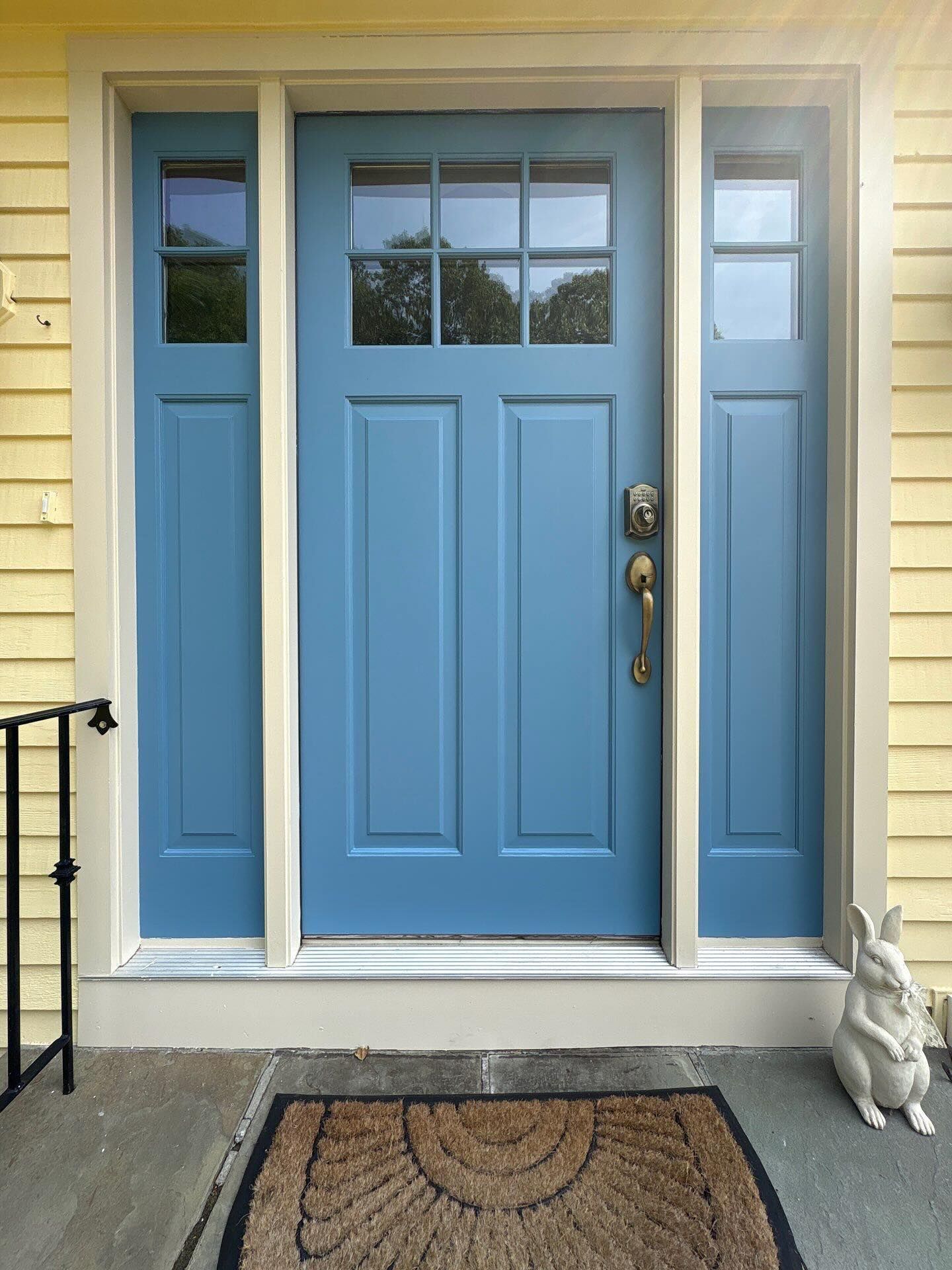 Blue front door with sidelights, off-white trim, on a yellow house. Doormat and rabbit statue.